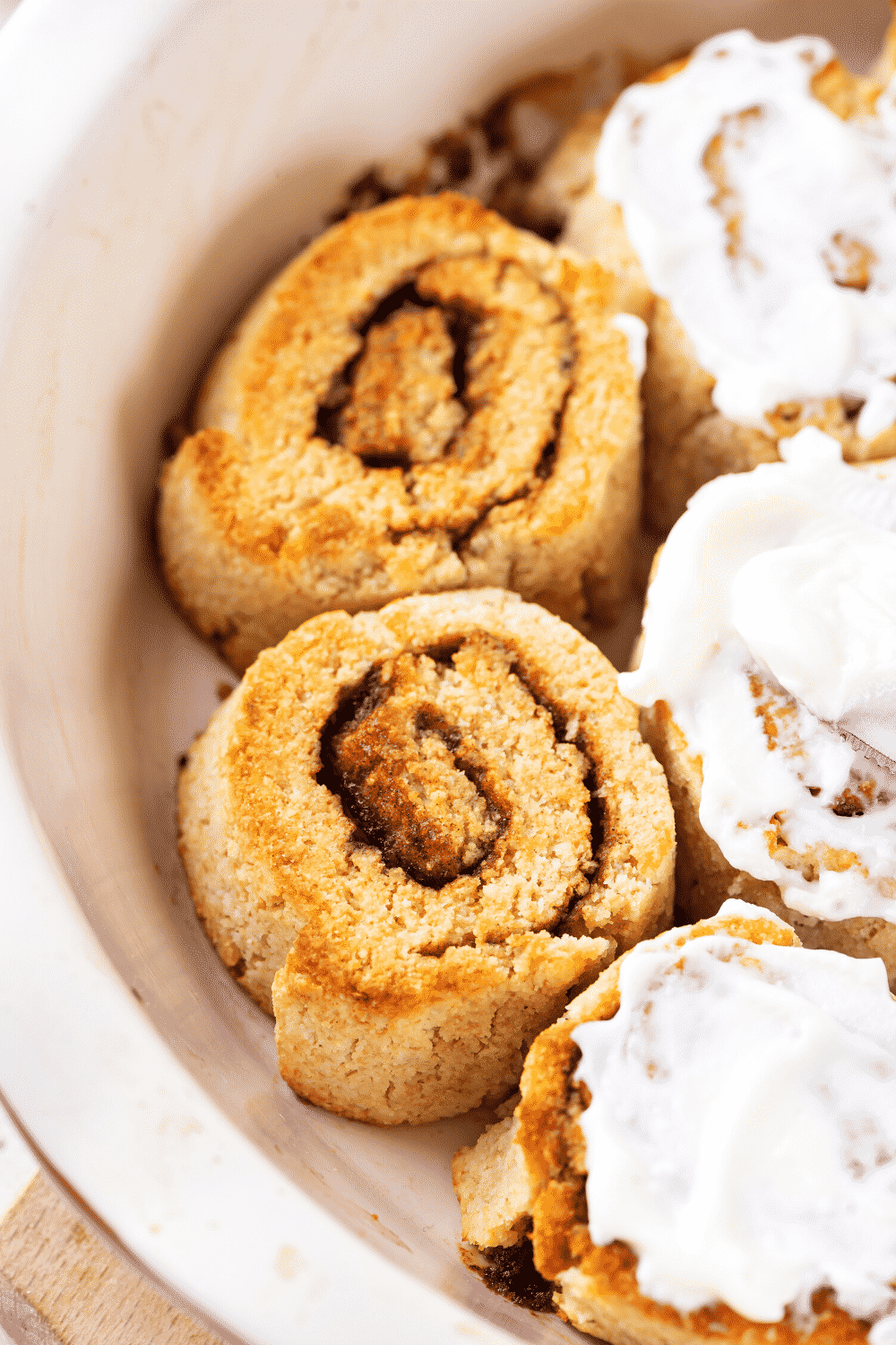 Two unfrosted cinnamon rolls in a white baking dish. There are three cinnamon rolls to the right of them with icing on top.