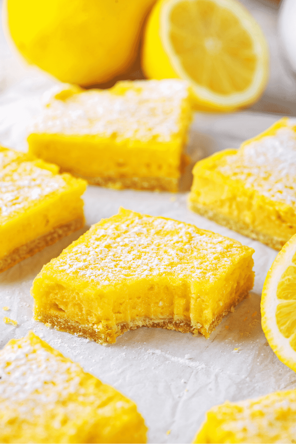 A few lemon bars on white parchment paper. The lemon bar in the middle has a bite out of the front. There are some lemons in the behind the lemon bars.