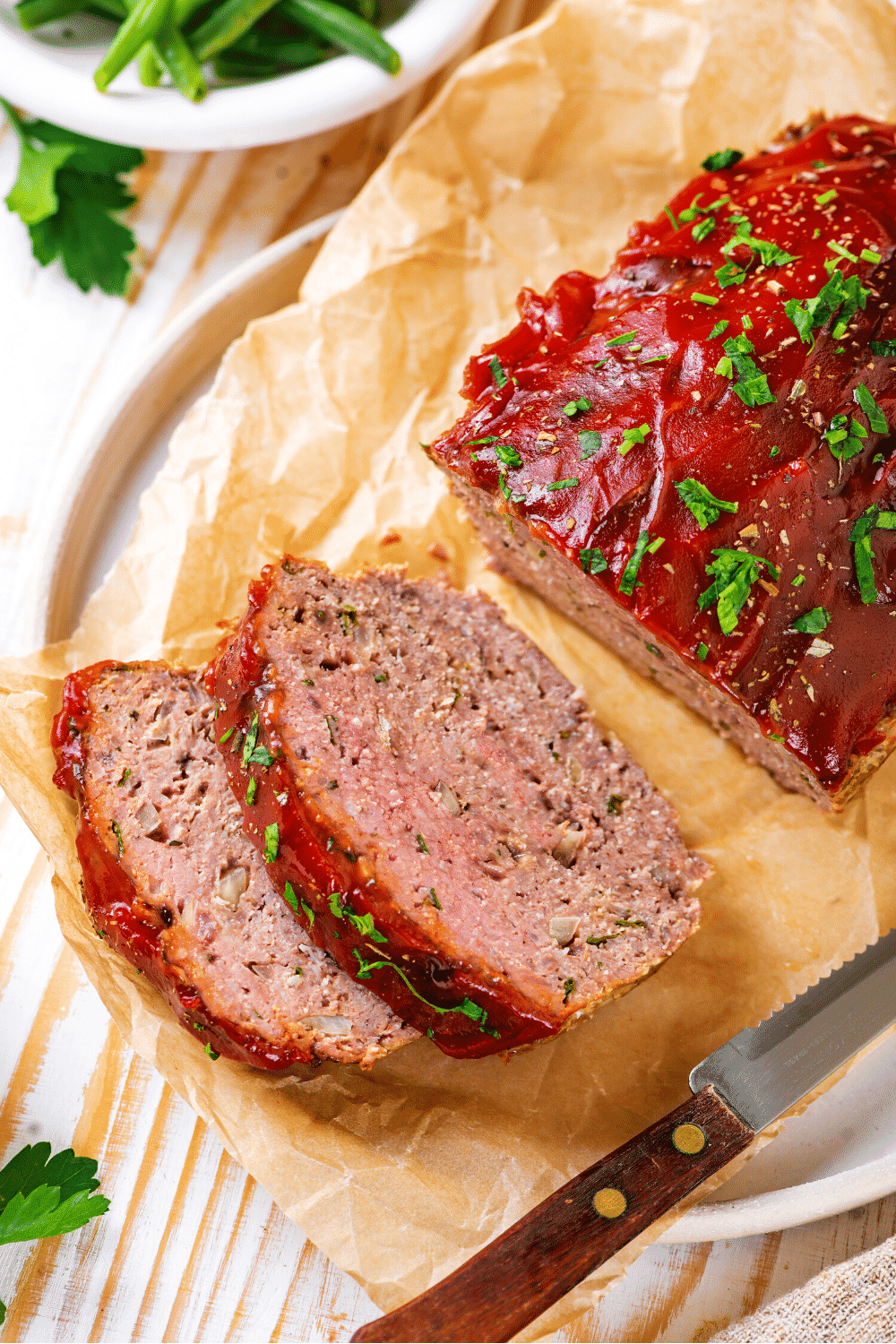 A whole meatloaf with two slices of meatloaf in front of it. The meatloaf is on a piece of parchment paper on a white plate.