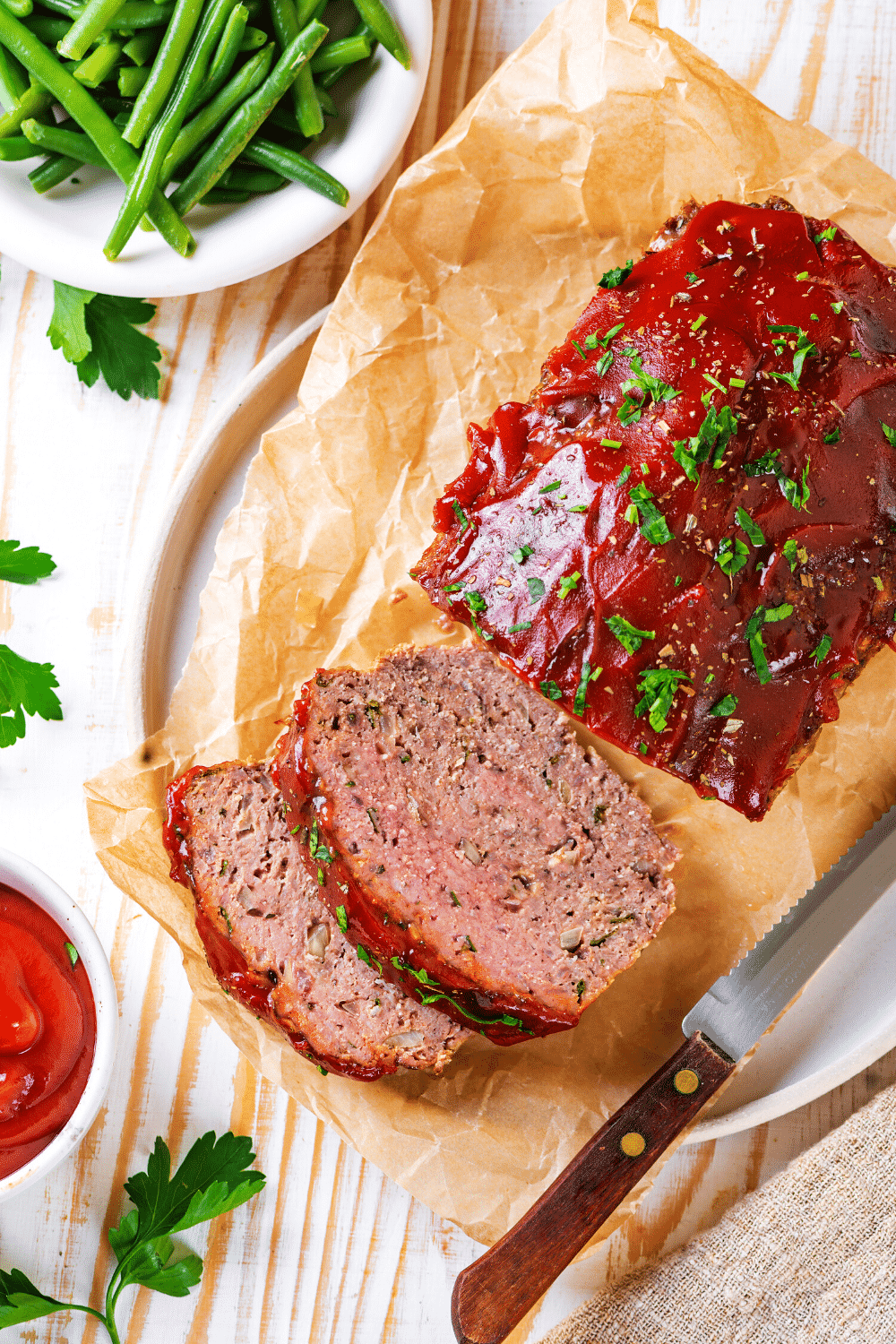 A whole meatloaf with two slices in front of it on a piece of parchment paper on a white plate.