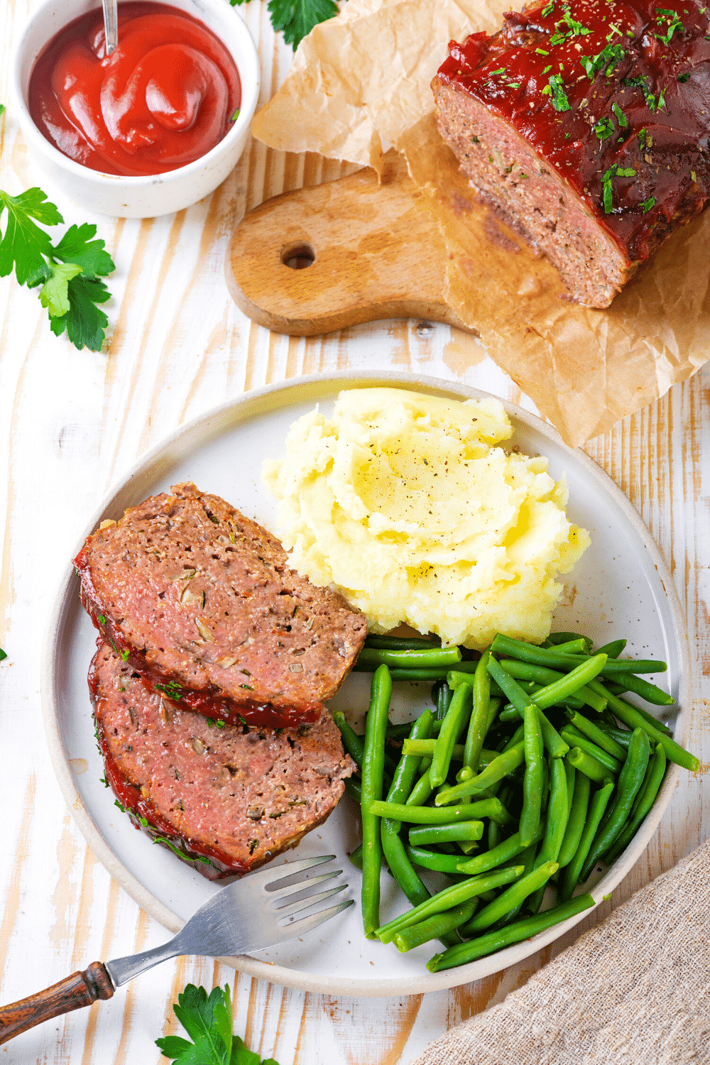 A white plate with two slices of meatloaf mashed potatoes and green beans. The whole meatloaf is behind the plate on a piece of white parchment paper on a wooden cutting board.