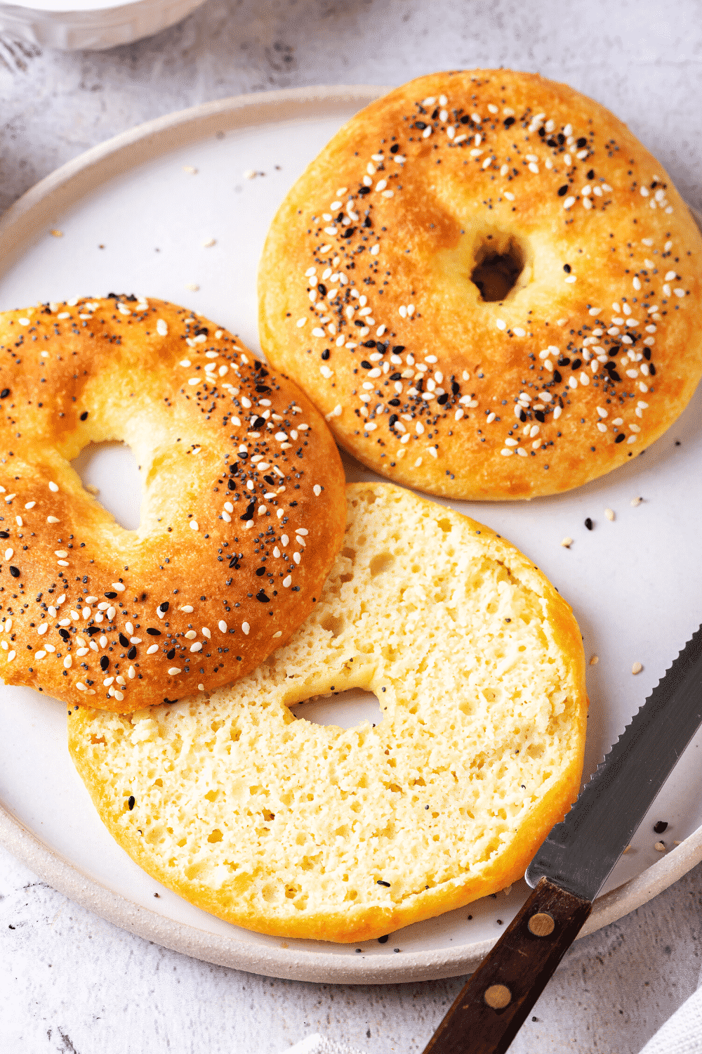 A white plate with a whole bagel and a sliced bagel. A serrated knife is next to the sliced bagel.