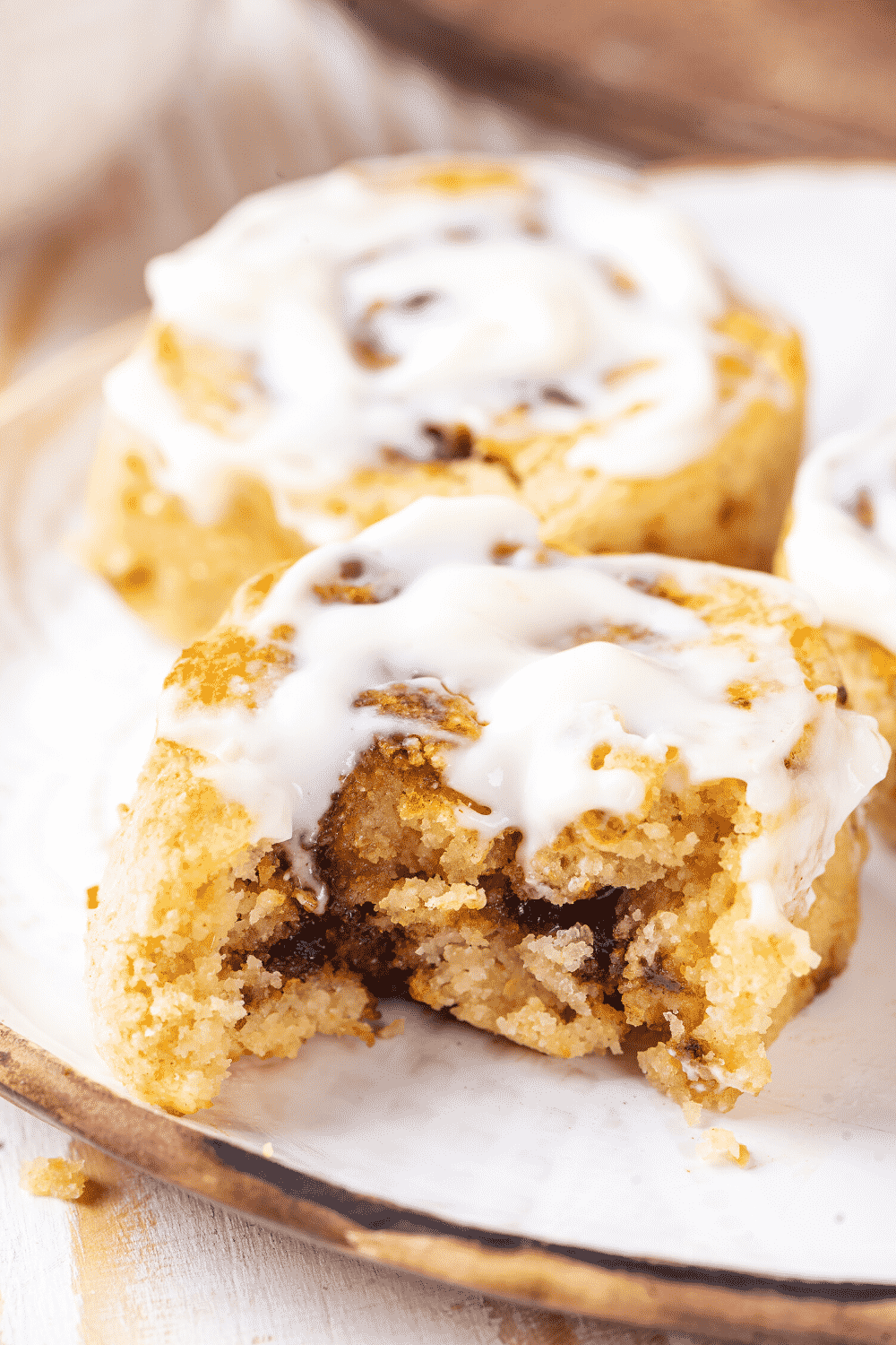 A cinnamon roll with a bite out of the front on a white plate. There is a whole cinnamon roll behind it