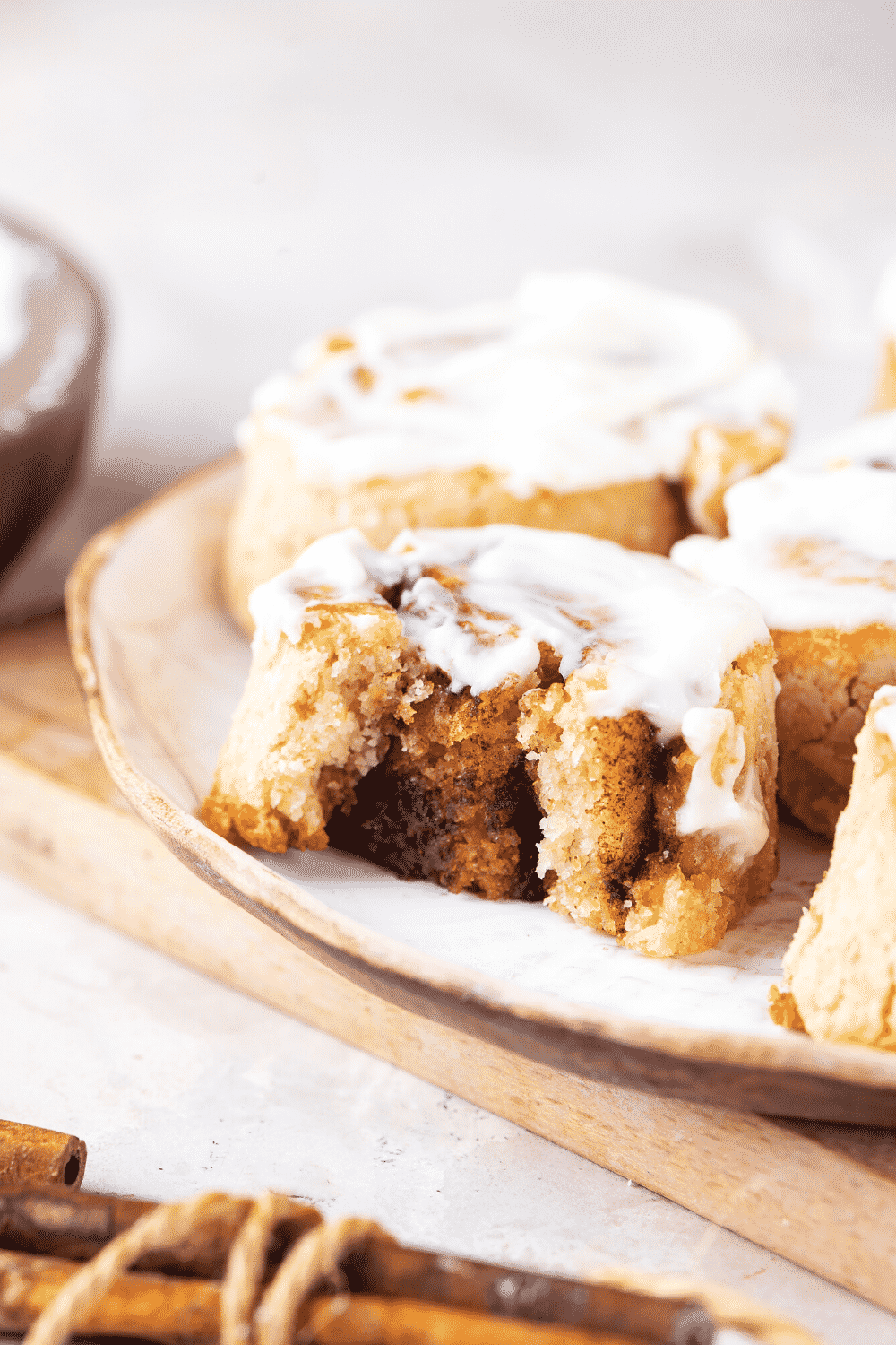 A white plate with a cinnamon roll with a bite out of the front. There is a whole cinnamon roll behind it and some more cinnamon rolls to the right of it.