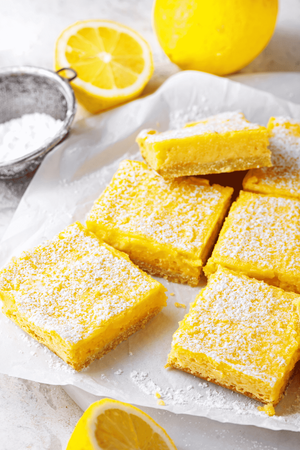 Two rows of three lemon bar squares on a piece of white parchment paper. There are some lemons behind the parchment paper and a bowl of powdered sugar.