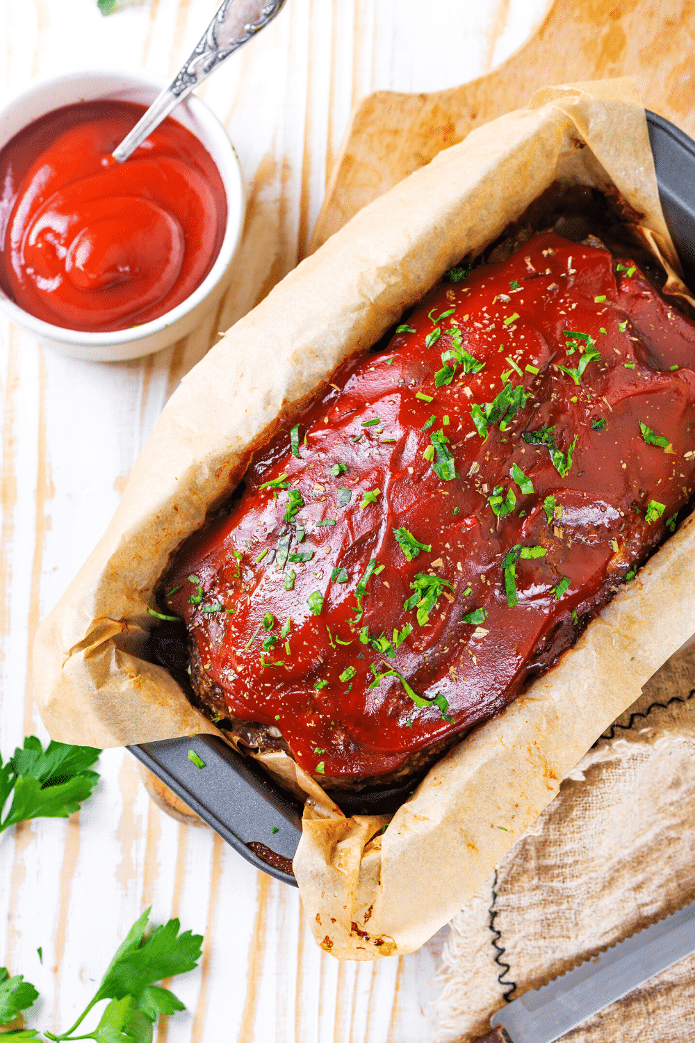 A meatloaf in a loaf pan with a piece of parchment paper underneath the meatloaf. A small cup of ketchup is to the back left of the meatloaf.