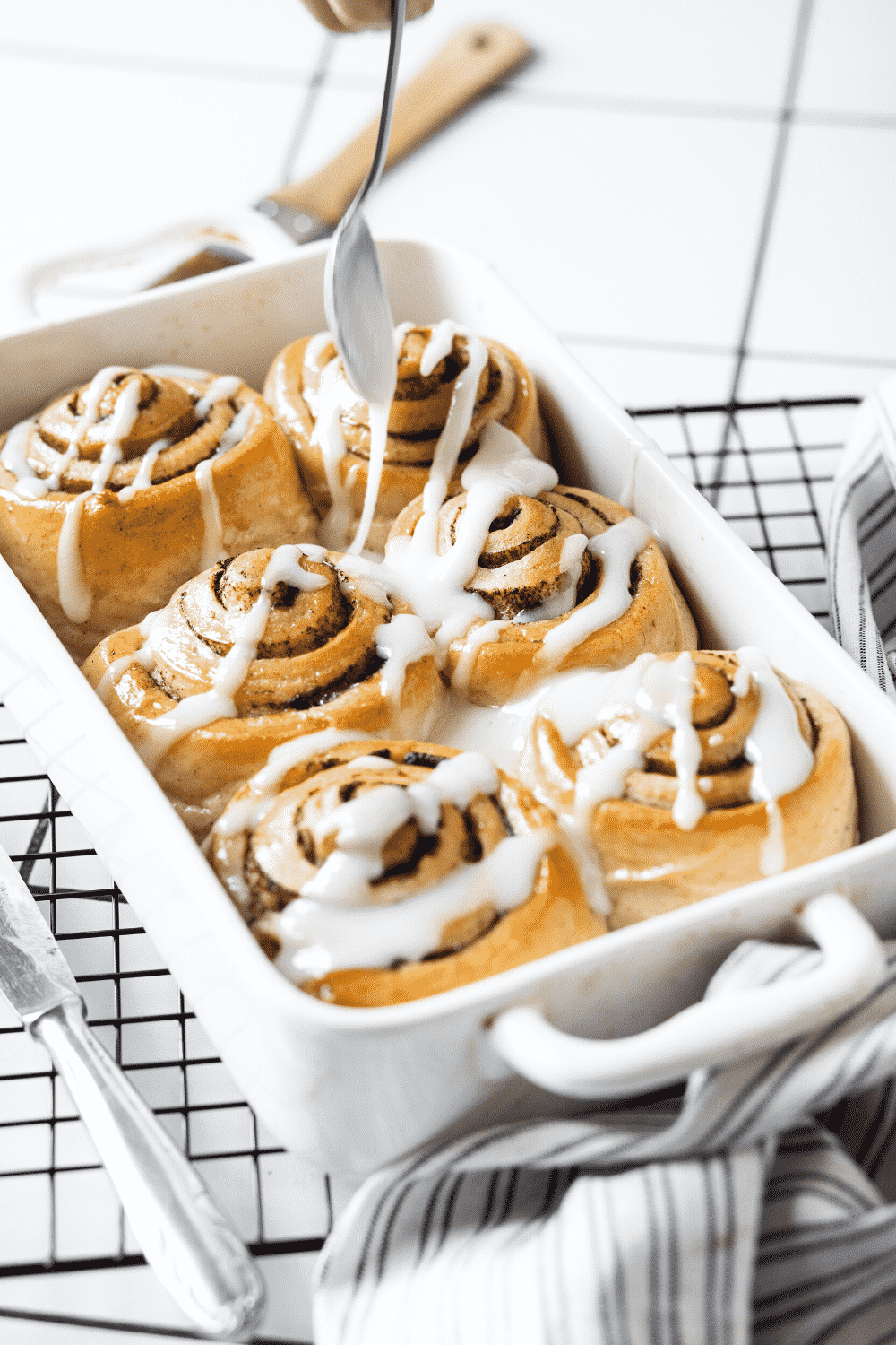 A square baking dish with six cinnamon rolls in it with a spoon hovering over the cinnamon rolls drizzle in icing on them.