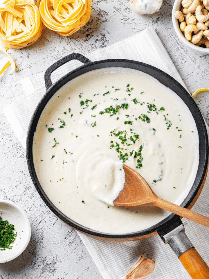 A black pan with Alfredo sauce in it. A wooden spoon is dipped in the Alfredo sauce with some sauce covering half of the surface of the spoon.