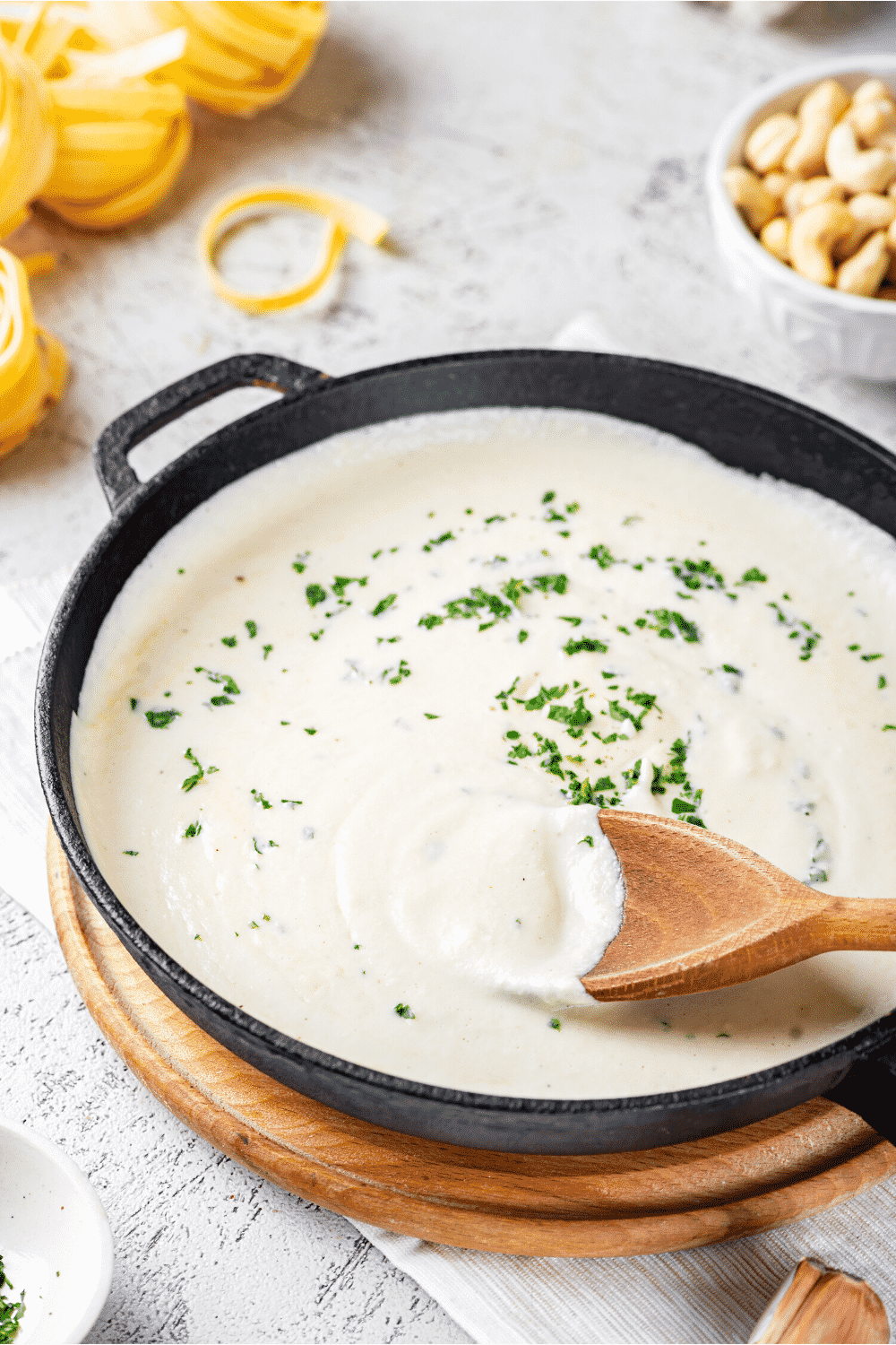 A black pan filled with dairy free Alfredo sauce. There is a wooden spoon in the Alfredo sauce with sauce covering half of the surface of the spoon.