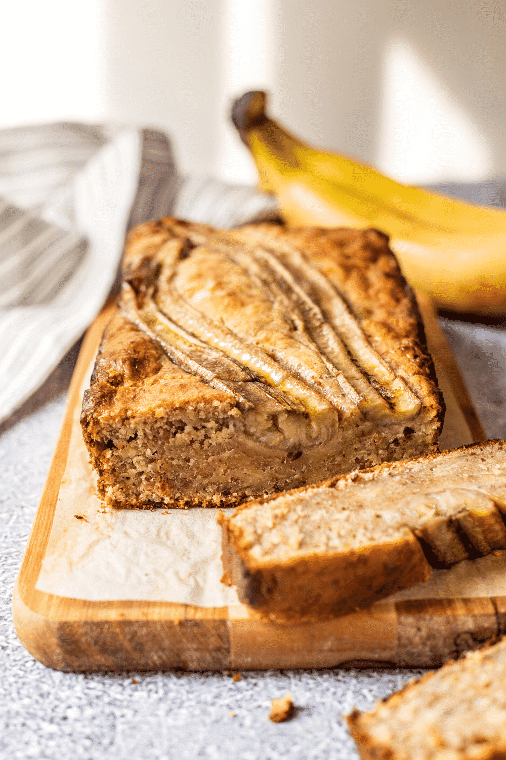 Loaf of banana bread on a piece of parchment paper on a wooden cutting board. There is a slice out of the loaf sitting in front of it and a bunch of bananas behind the loaf.