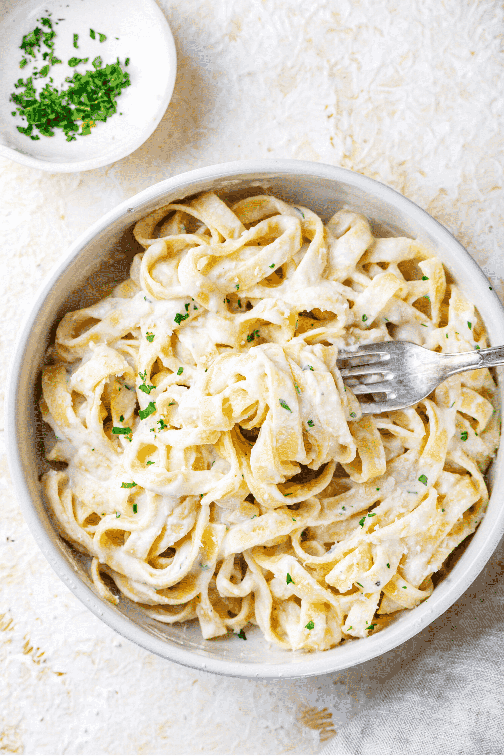 A white bowl filled with fettuccine Alfredo. A fork has fettuccine noodles inner twine in the prongs and is in the bowl.