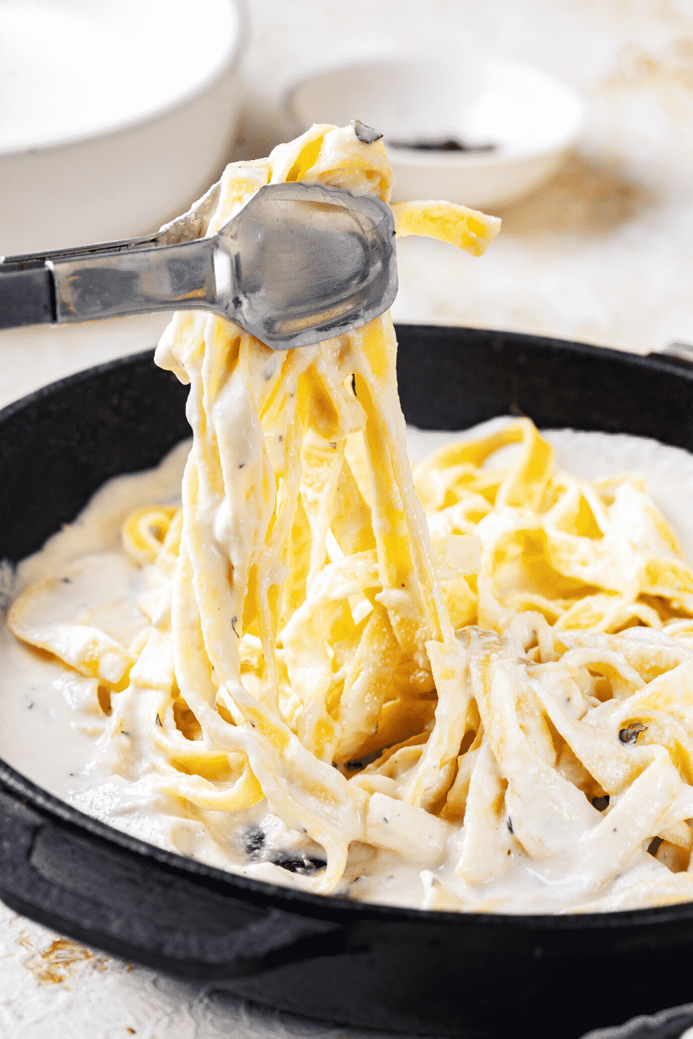 A black pan filled with fettuccine Alfredo. Tongs are hovering over the pan with fettuccine noodles between them draped down into the pan.