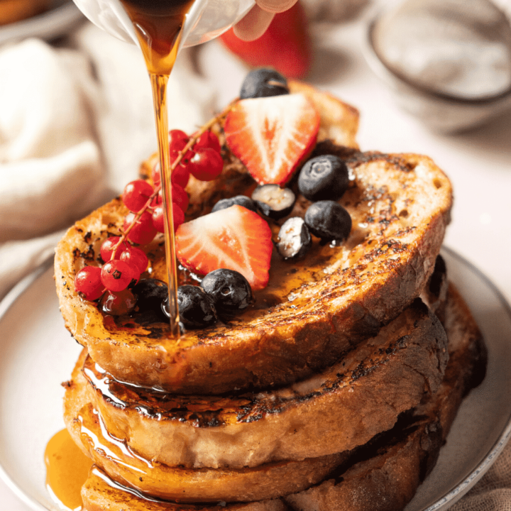 Four slices of vegan French toast stacked on top of one another on a white plate. Fresh fruit is on the top slice of French toast and maple syrup is being poured over the stack.