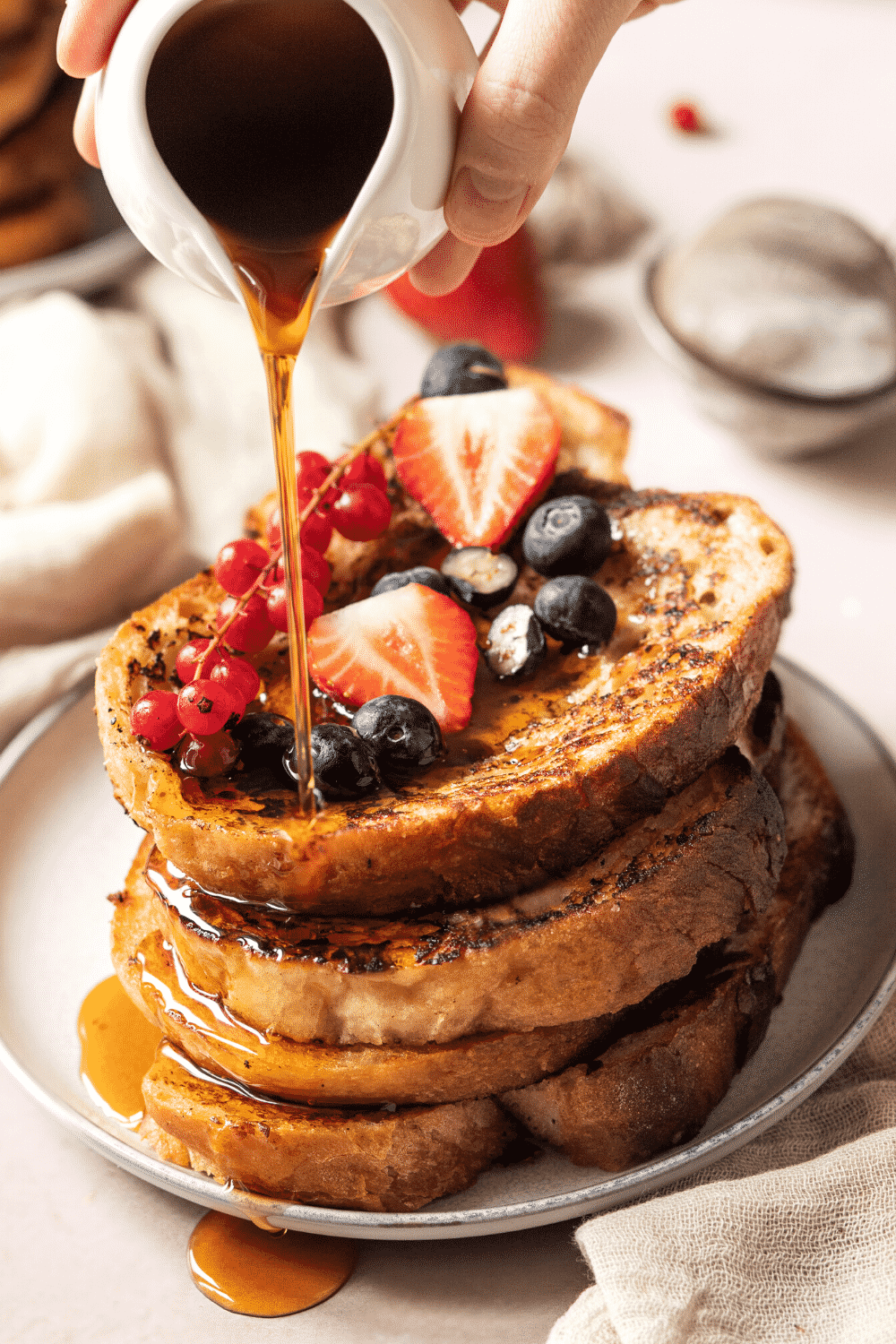 Four slices of French toast stacked on top of one another on a white plate. Fresh fruit is on the top slice of French toast and maple syrup is being poured over the stack.