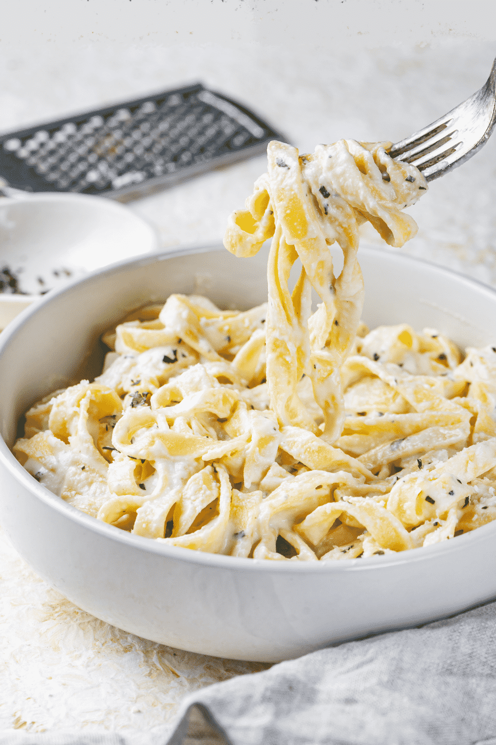 A white bowl filled with fettuccine Alfredo. A fork is hovering over the ball and has fettuccine noodles in the prongs and touching the rest of the noodles in the bowl.