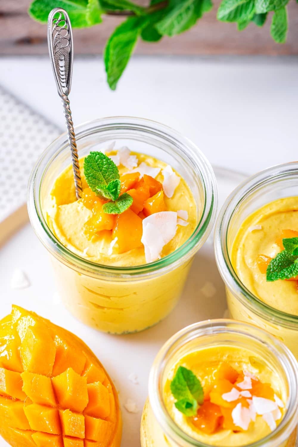 A cup of mango mousse on a white plate. There is part of a cup to the right of it and a small cup in front of it both filled with mango mousse.