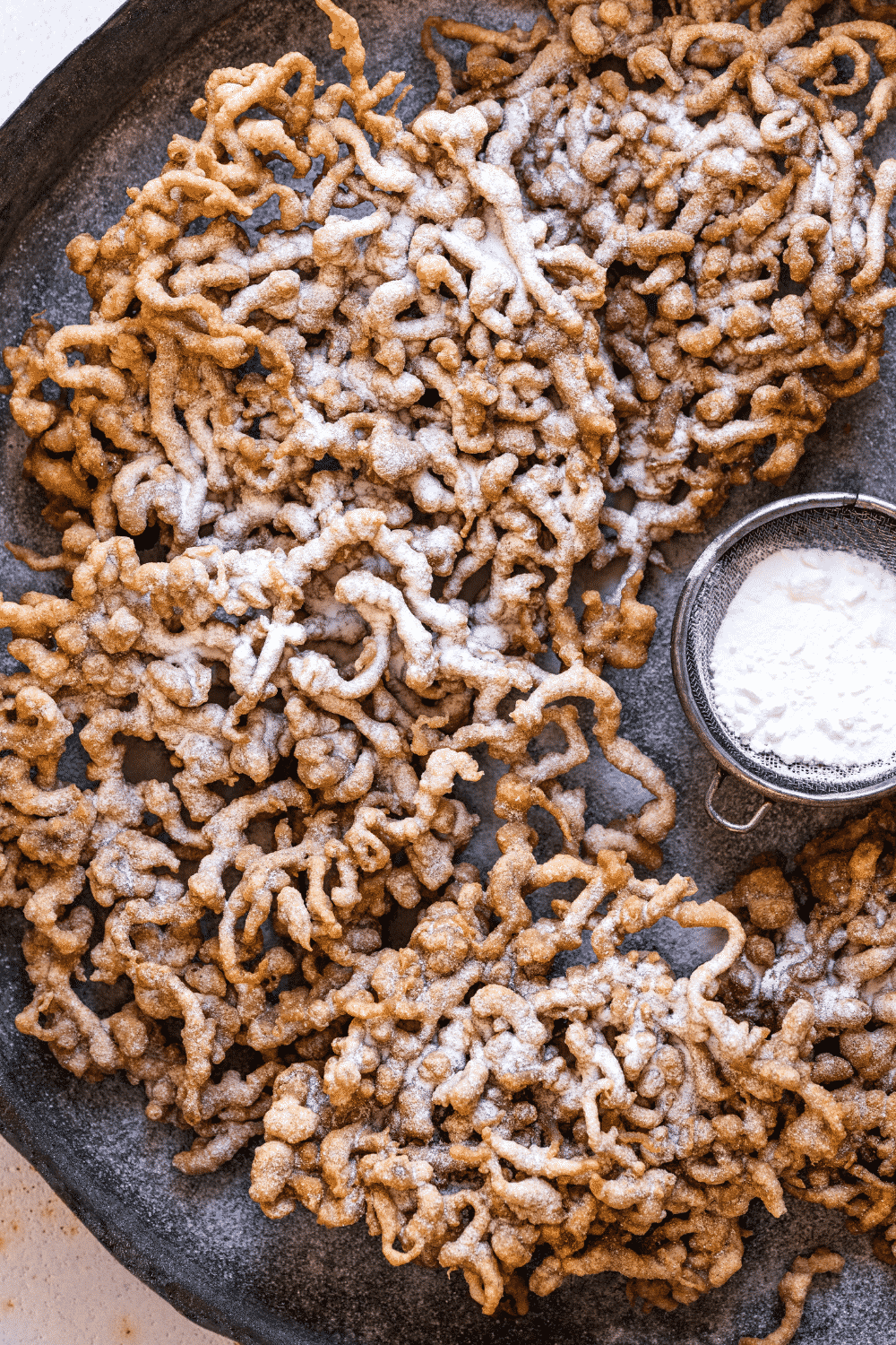 A bunch of funnel cakes on a black plate. A small sifter filled with powdered sugar is to the right of the funnel cakes.
