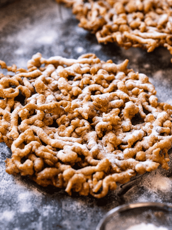 A funnel cake and a black table with another funnel cake behind it. The table is covered in powdered sugar and there is a small sifter of powdered sugar at the front of the funnel cake.