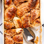 A white casserole dish filled with bread pudding. A chunk of bread putting is missing from the front right of the casserole dish and there is a metal spoon in its place.