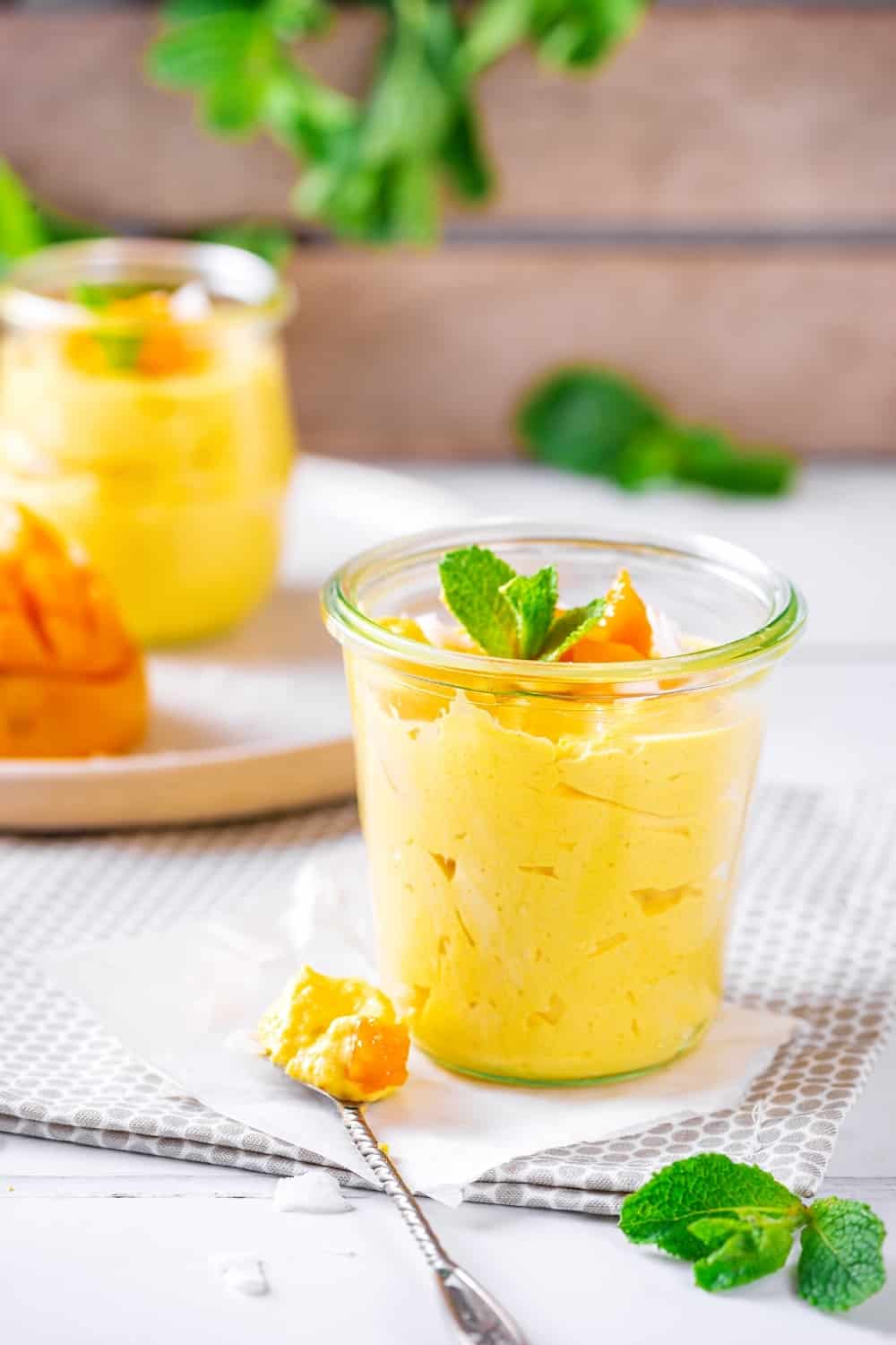 A glass cup filled with mango mousse on a napkin set on a tablecloth on the white table. Hey spoon with a scoop of mango mousse on it is on the napkin.