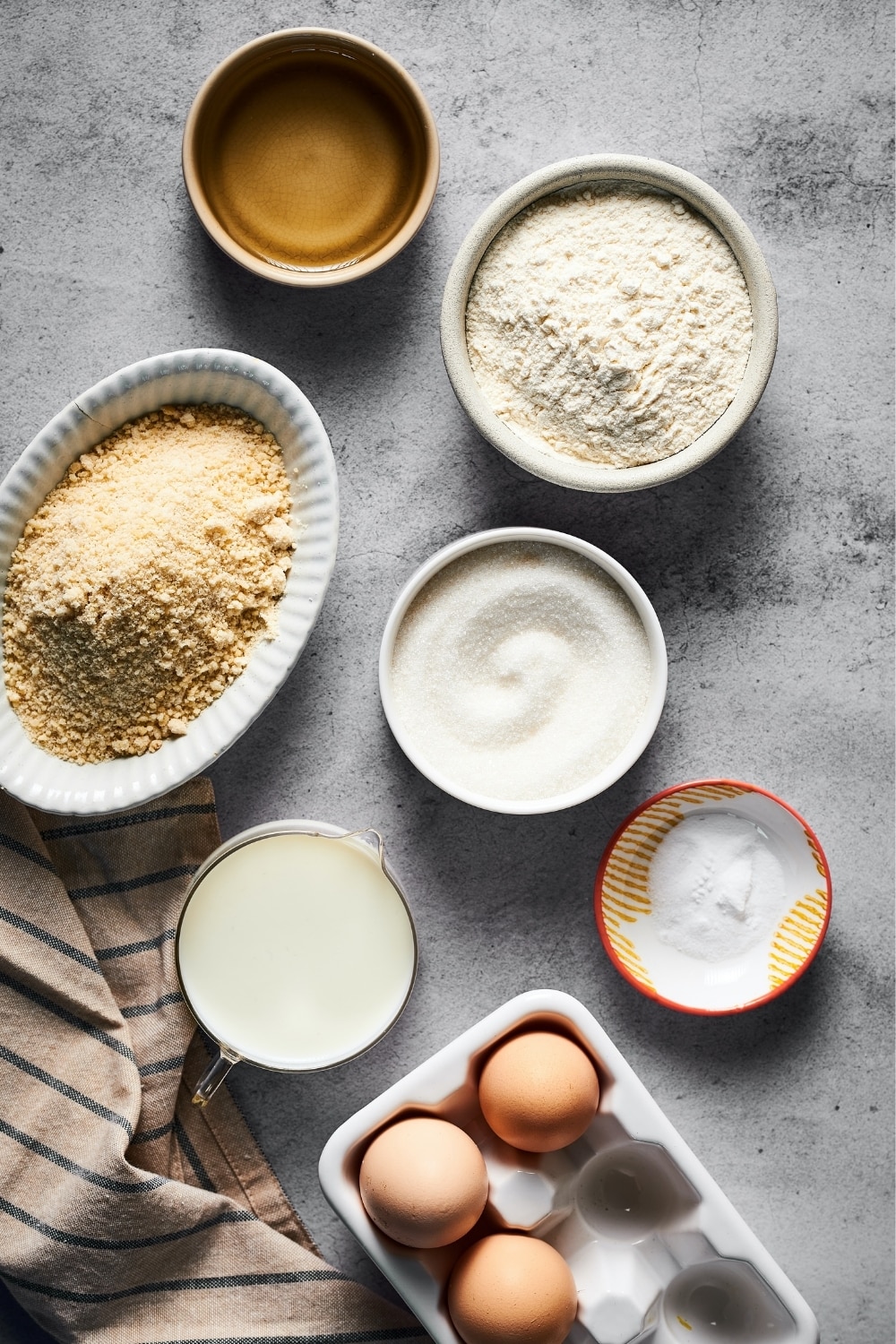 A small bowl of sugar, a small bowl of vegetable oil, a medium bowl of flour, an oval bowl of almond flour, and a measuring cup of buttermilk on a gray counter. A carton of eggs is in front of all the bowls.