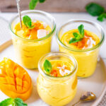 3 glass cups of mango mousse on a white plate. There is diced mango and coconut flakes on top of the mousse.