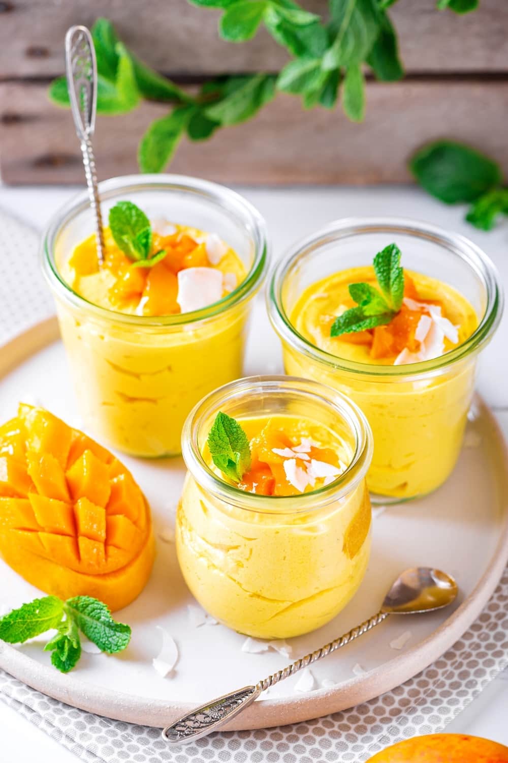 3 glass cups of mango mousse on a white plate. There is diced mango and coconut flakes on top of the mousse.