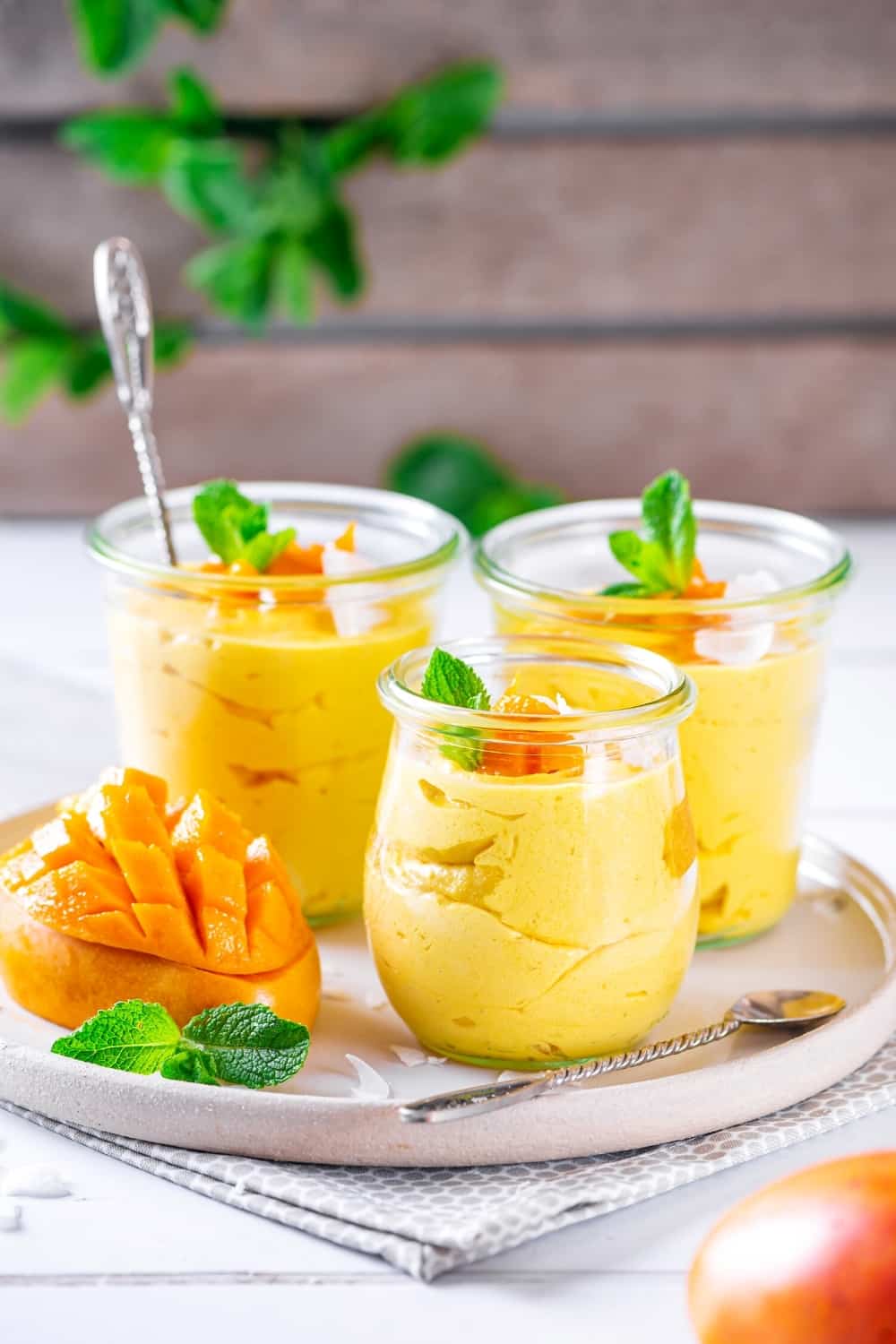3 glass cups of mango mousse on a white plate. There is a sliced mango to the left of the cups on the plate.