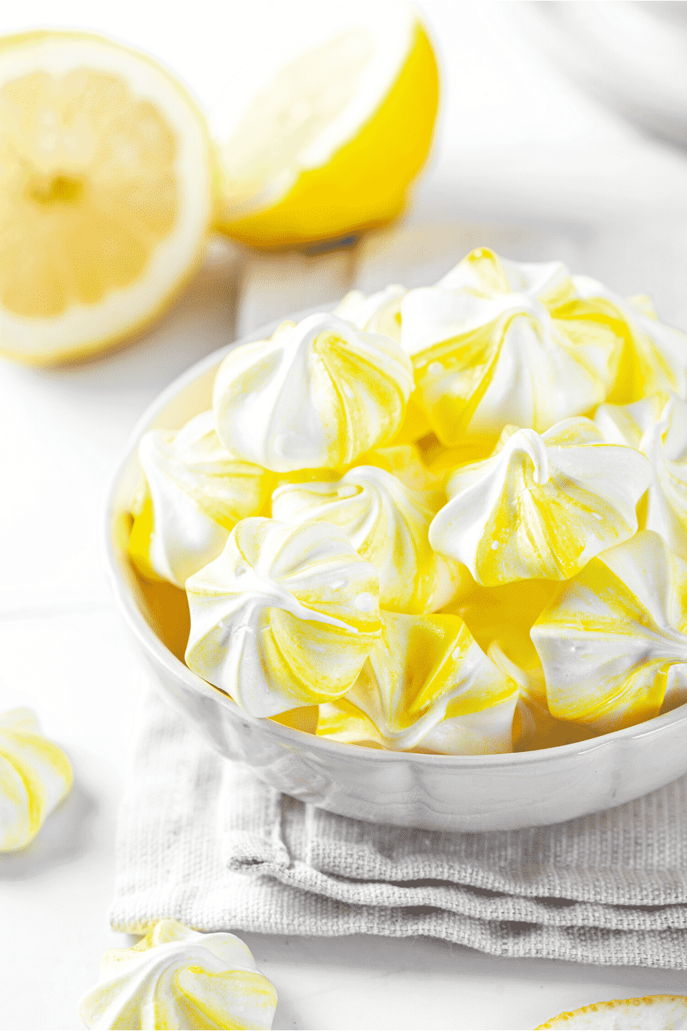 A white bowl filled with lemon meringue cookies. The ball is on the table cloth and a white counter with some lemons behind it.
