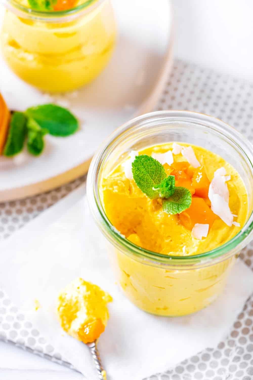 An overhead view of a glass cup of mango mousse white napkin on the table cloth. The boss has a scoop out of it and there is a spoon on the napkin with the scoop on it.