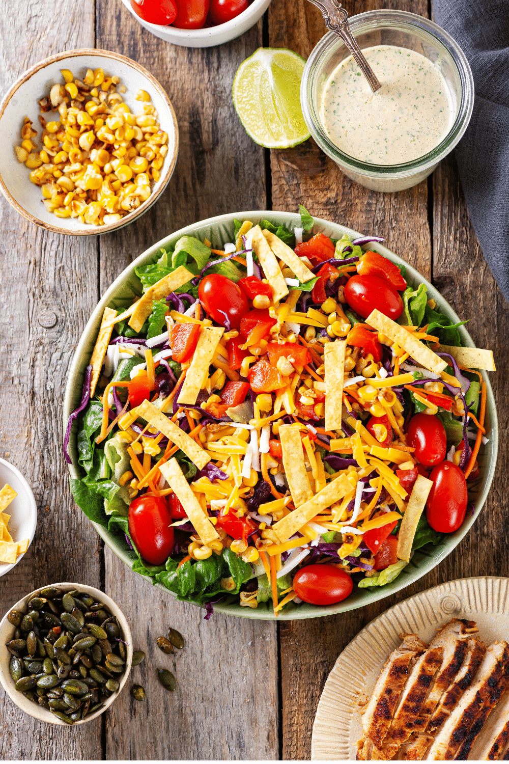 Is southwest salad in a white bowl. There is a small white cup of chili lime pepitas in front of it, a small white bowl of corn behind it, and a small glass jar of creamy salsa dressing behind it.