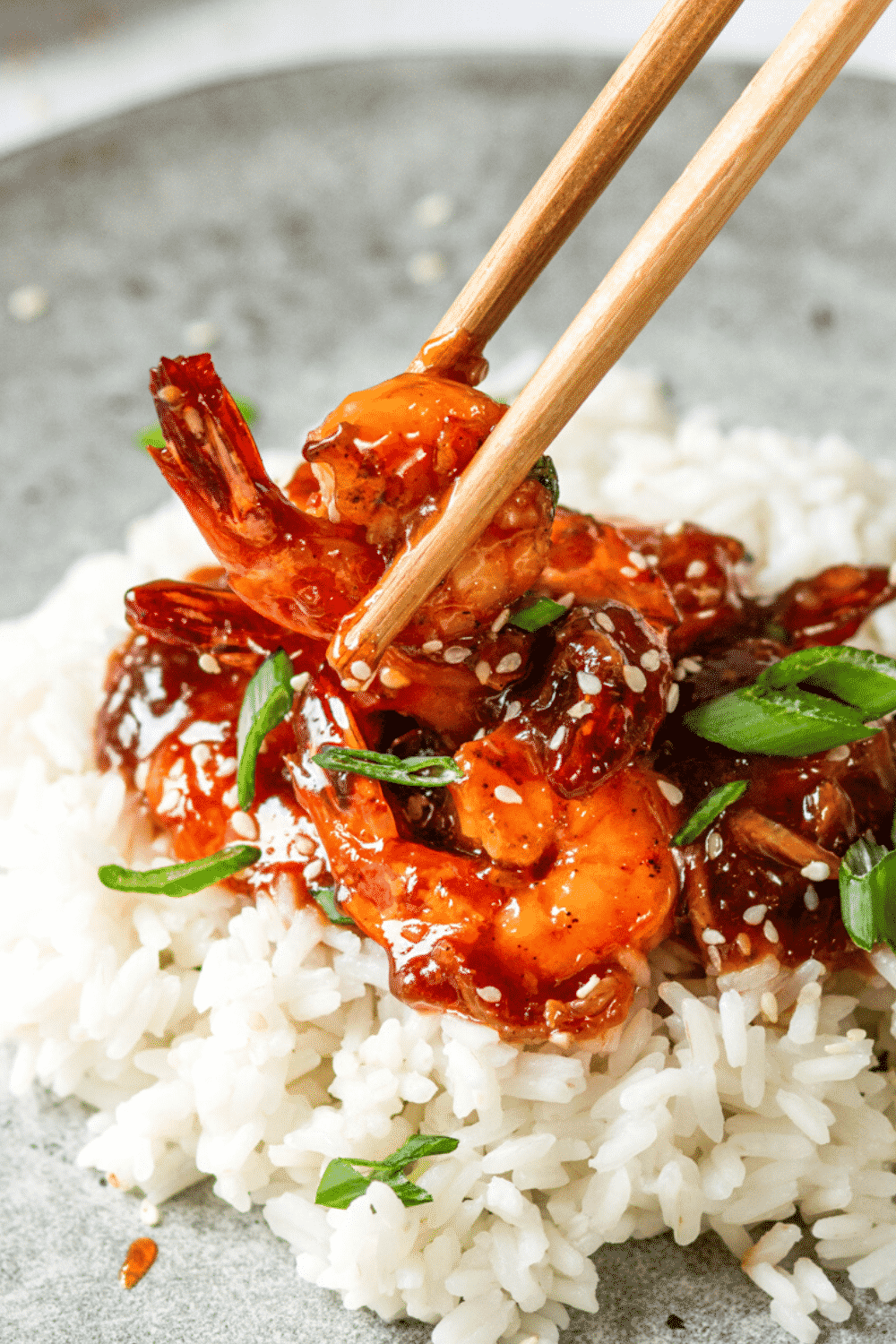 A piece of sweet and sour shrimp being held by Chopstix over a few sweet-and-sour shrimp and a bag of white rice on a plate.
