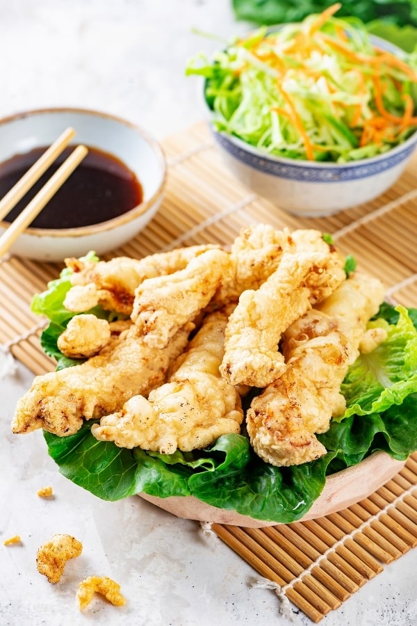 A wooden bowl filled with lettuce and pieces of chicken tempura on top. Behind it is a small bowl of soy sauce and next to that small bowl of shredded cabbage all on a wooden placemat.