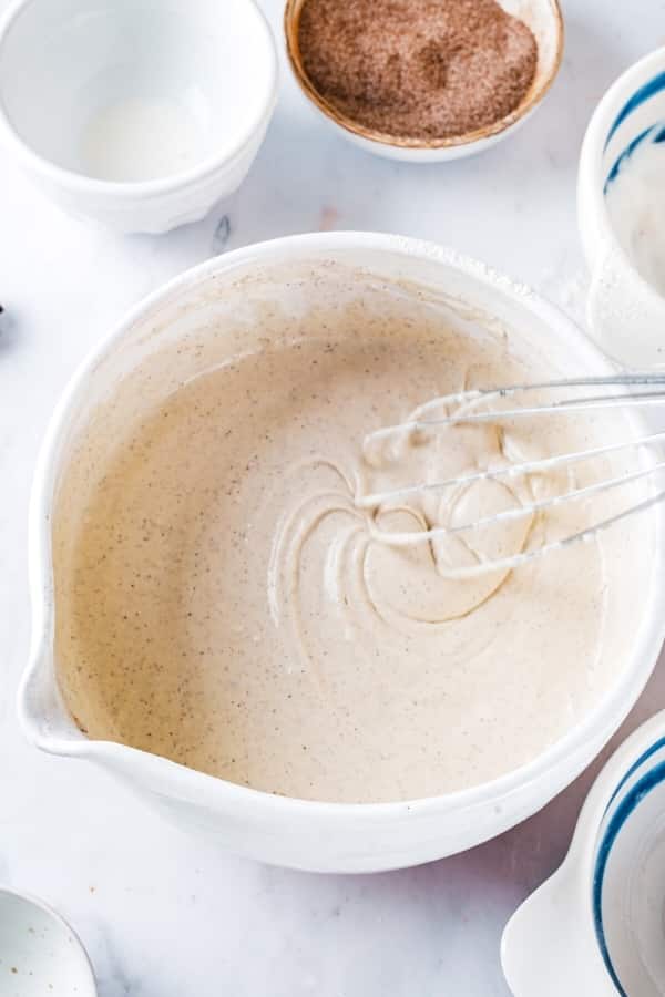 A white mixing bowl with churro pancake batter in it. There is a whisk in the bowl and behind the ball is a small bowl of cinnamon sugar.