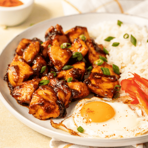 Chicken tocino, a fried egg, part of sliced red bell peppers, and some white rice, on a white plate. The plate is on a white and gold checkered tablecloth on a white counter.