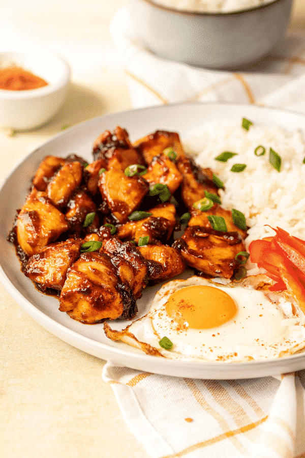 Chicken tocino, a fried egg, part of sliced red bell peppers, and some white rice, on a white plate. The plate is on a white and gold checkered tablecloth on a white counter.