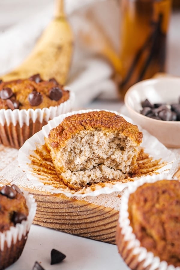A almond flour banana muffin with the front cut out of it on a muffin wrapper. There is part of a chocolate chip banana muffin behind it and parts of two muffins in front of it.