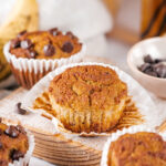 An almond flour banana muffin on top of a muffin wrapper on a wooden cutting board. There is a chocolate chip banana muffin behind it and parts of two banana muffins in front of the cutting board.