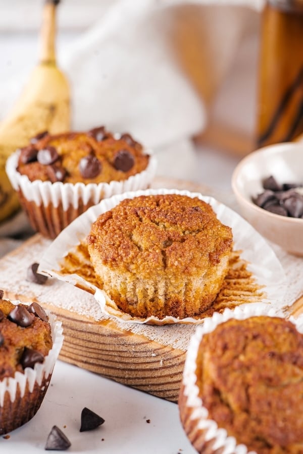 An almond flour banana muffin on top of a muffin wrapper on a wooden cutting board. There is a chocolate chip banana muffin behind it and parts of two banana muffins in front of the cutting board.