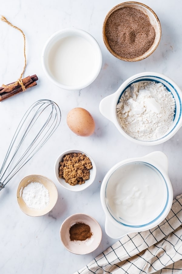 A small bowl of cinnamon sugar, a small bowl of milk next to it, a whisk and egg in front of it, a medium bowl of flour next to the egg, in front of that a medium bowl of yogurt, and to the left a small bowl of cinnamon, a small bowl baking powder, and a small bowl of brown sugar. Everything is sitting on a white counter.