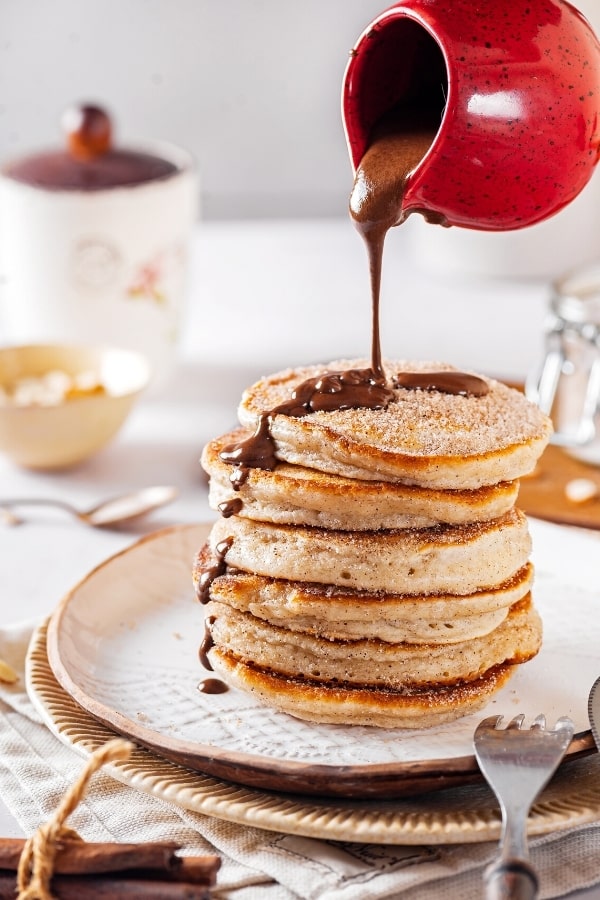 Stack of six churro pancakes on a white plate. There is a red cup pouring chocolate sauce on top of the pancakes.