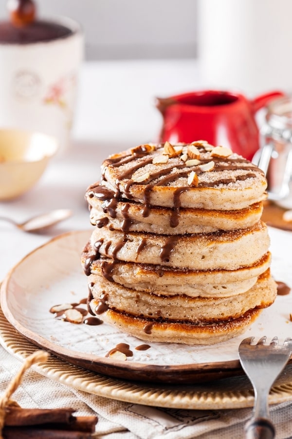 Five churro pancakes with chocolate sauce drizzled on top stacked on a white plate.