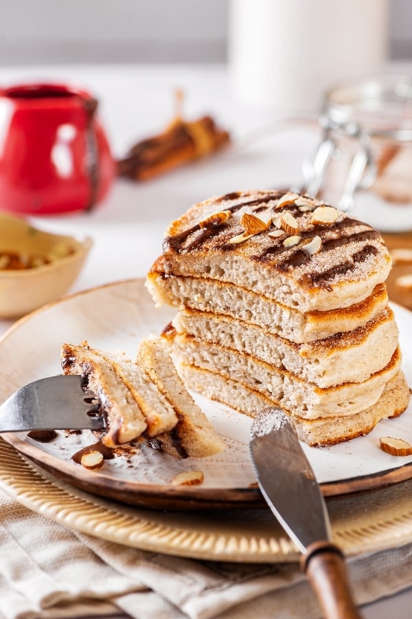 White plate with a stack of six churro pancakes on it. The top pancake has some chocolate sauce on top and there is a fork at the front of the plate with three pieces of pancake on the prongs.