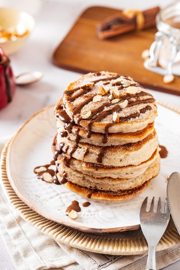 A stack of six churro pancakes with chocolate sauce drizzled on top on a white plate. There is part of a fork and knife at the front of the plate and behind the plate is a wooden cutting board with a bundle of cinnamon on it.