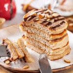 A stack of six churro pancakes on a white plate with a piece cut out of the front of all the pancakes. There are three pieces of pancake on the prongs of a fork at the front edge of the plate.