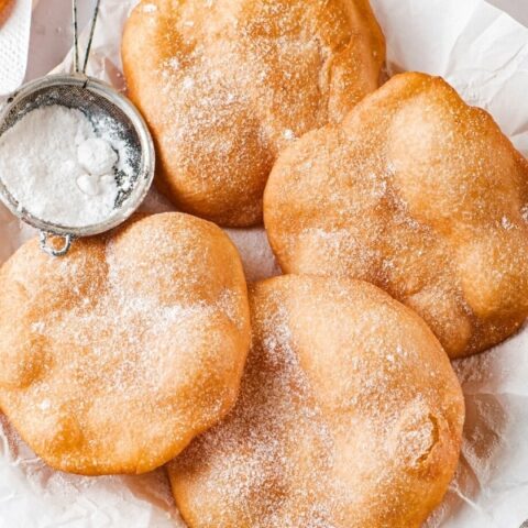 Four pieces of fried dough powdered sugar on them on a piece of parchment paper. A small sifter is at the left side of the parchment paper between two pieces of fried dough.