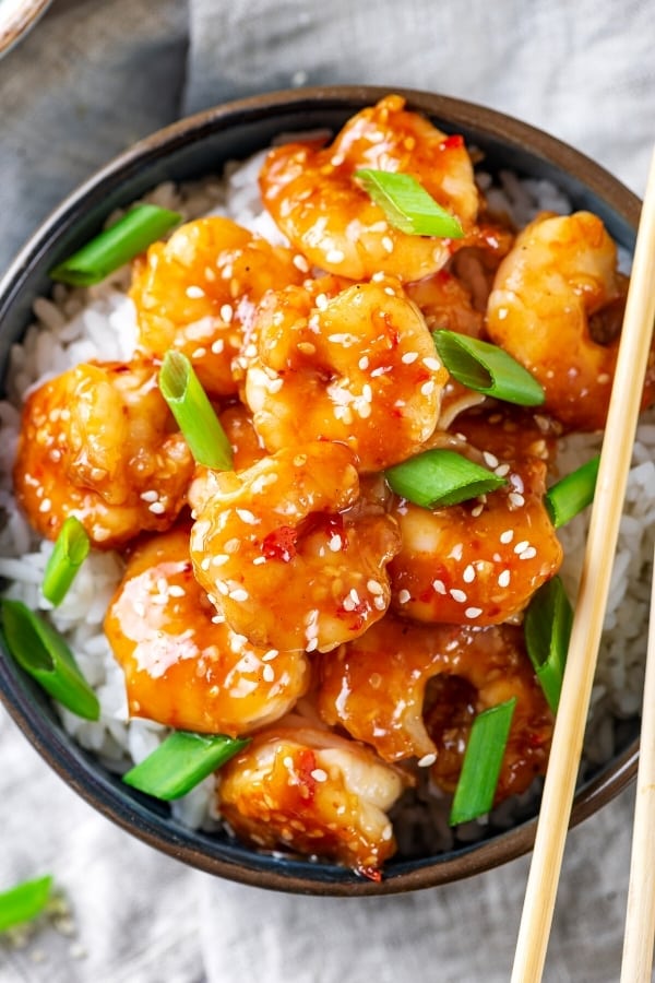 A bowl filled with a bunch of general Tso's shrimp and a few green onions on top of white rice. Part of one chopstick is on the edge of the bowl.