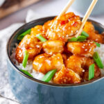 A blue bowl with white rice in it and General tsos shrimp and green onions on top. There is a set of chopsticks hovering over the bowl with a piece of shrimp between them.
