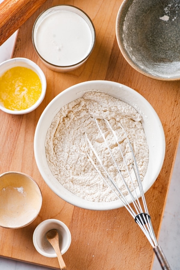 A large white bowl of flour and baking powder with a whisk in it. There's a small bowl of melted butter and a medium sized bowl of milk behind it. Everything is on a wooden cutting board.