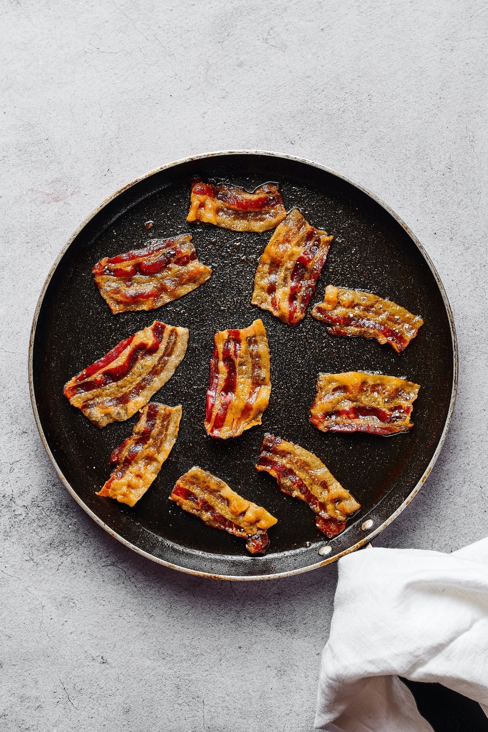 A black pan with 10 pieces of crispy bacon on it. The pan is on a grey counter and there is a white tablecloth over the handle.